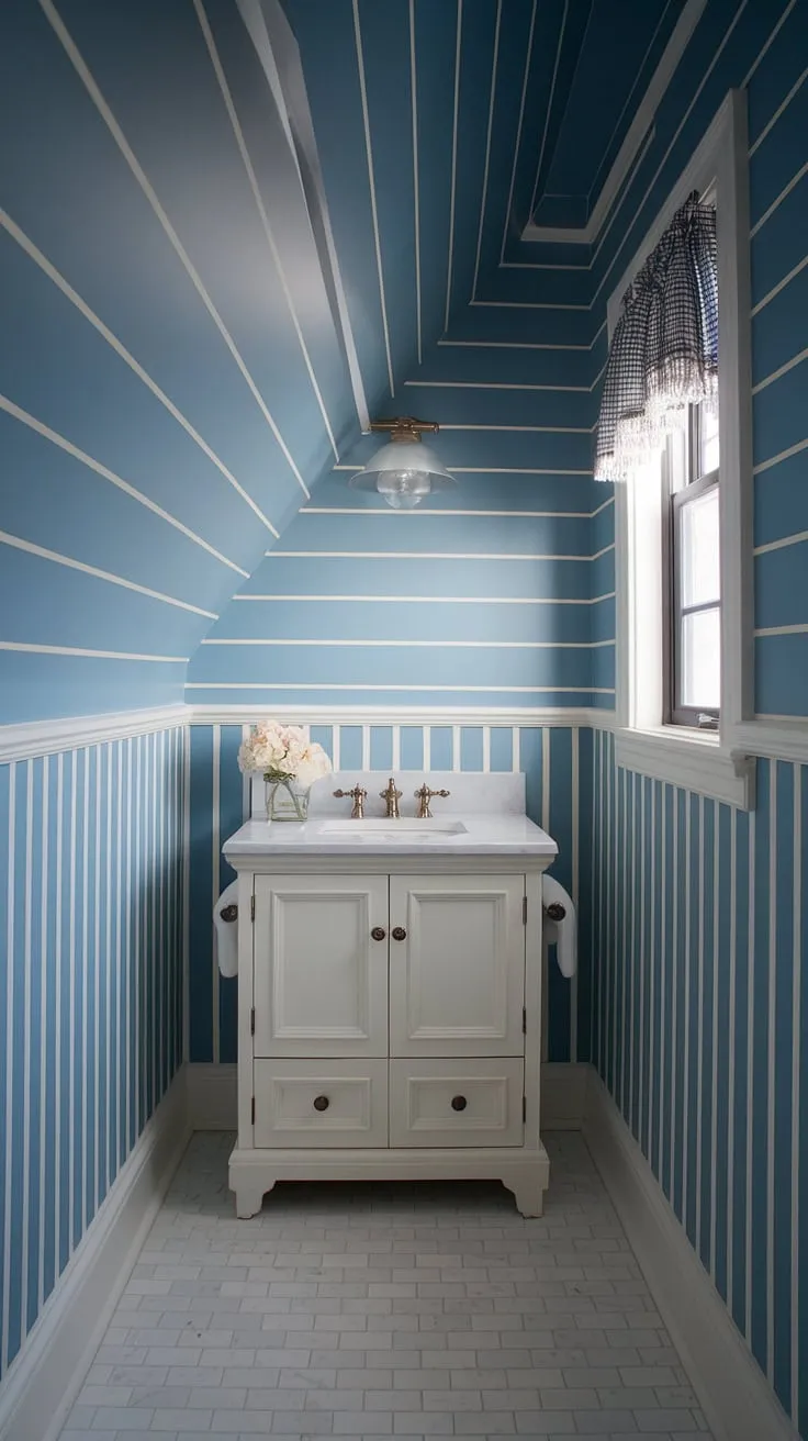 A serene coastal powder room with a slanted ceiling, featuring a stunning blue wallpaper bathroom.  The blue and white wallpaper bathroom creates a bright and airy feel, enhanced by the Serena &#038; Lily bathroom vanity and crisp white tile floor.  A perfect example of a stylish and functional powder room.