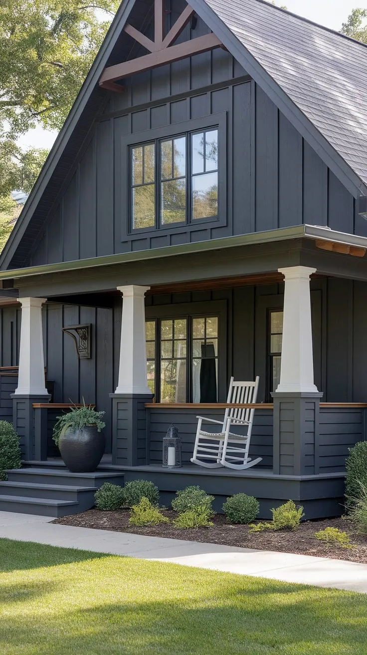 This dark gray modern farmhouse boasts a captivating blend of architectural styles. A thoughtfully designed front porch, complete with white columns and a rocking chair, invites relaxation and offers a picturesque view of the lush green landscape. A true testament to timeless design.