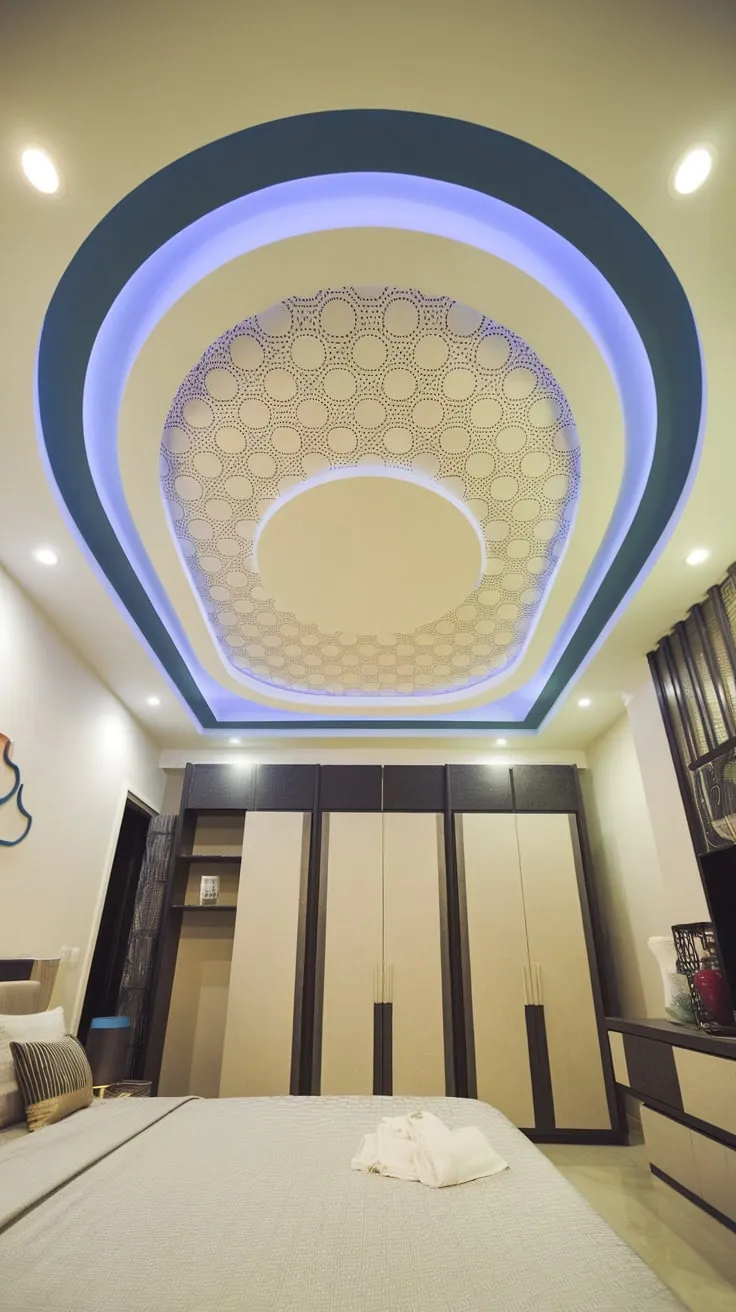 A modern bedroom's gypsum board false ceiling features a curved pop design with a circular blue-bordered pattern and LED lighting, enhancing the room's ambiance