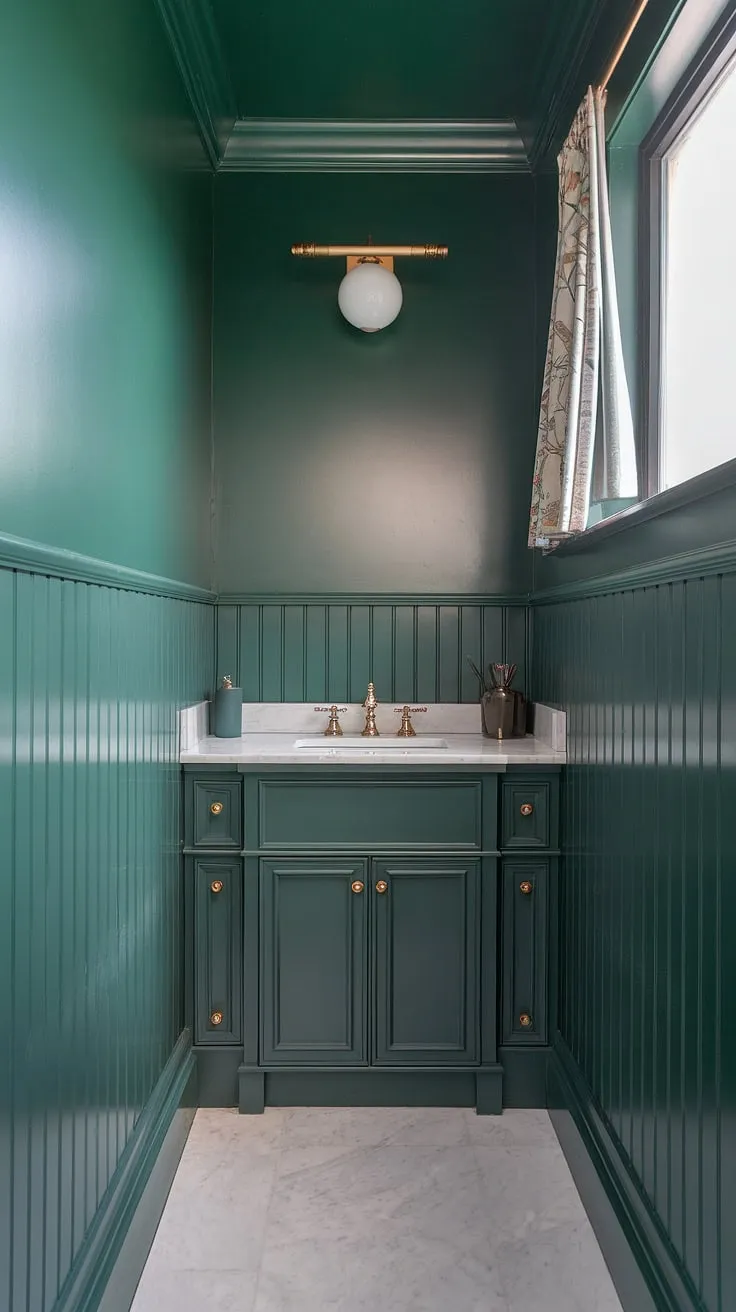 A charming small master bath in a serene olive green and white bathroom scheme. The green wall bathroom decor features striking green wainscoting that runs the height of the room, paired with a dark green vanity. The white marble floor brightens the space, while a gold light fixture adds a touch of glamour. This small dark green bathroom showcases clever green vanity powder room ideas, proving that a muted green bathroom can be both stylish and functional. This green full bathroom is a perfect example of small master bath green design.
