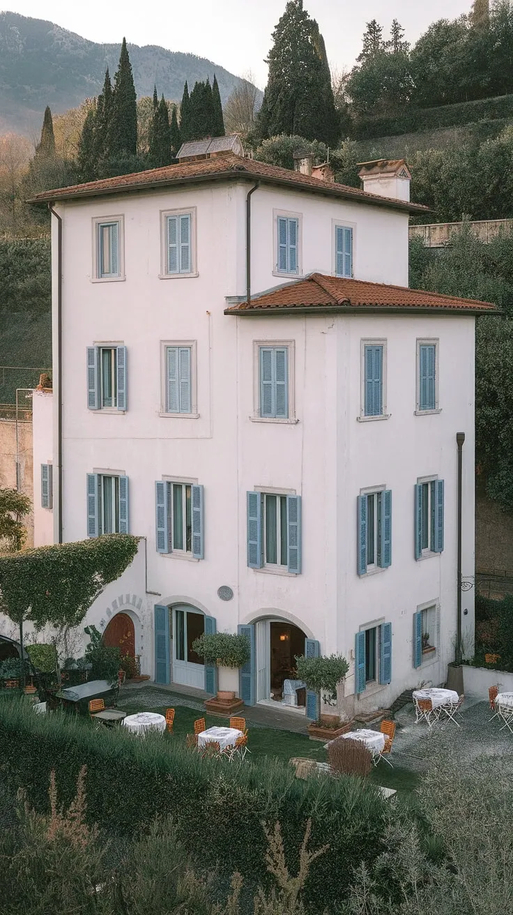 A charming Italian villa, complete with arched windows and outdoor seating, sits nestled amongst lush greenery and majestic mountains.