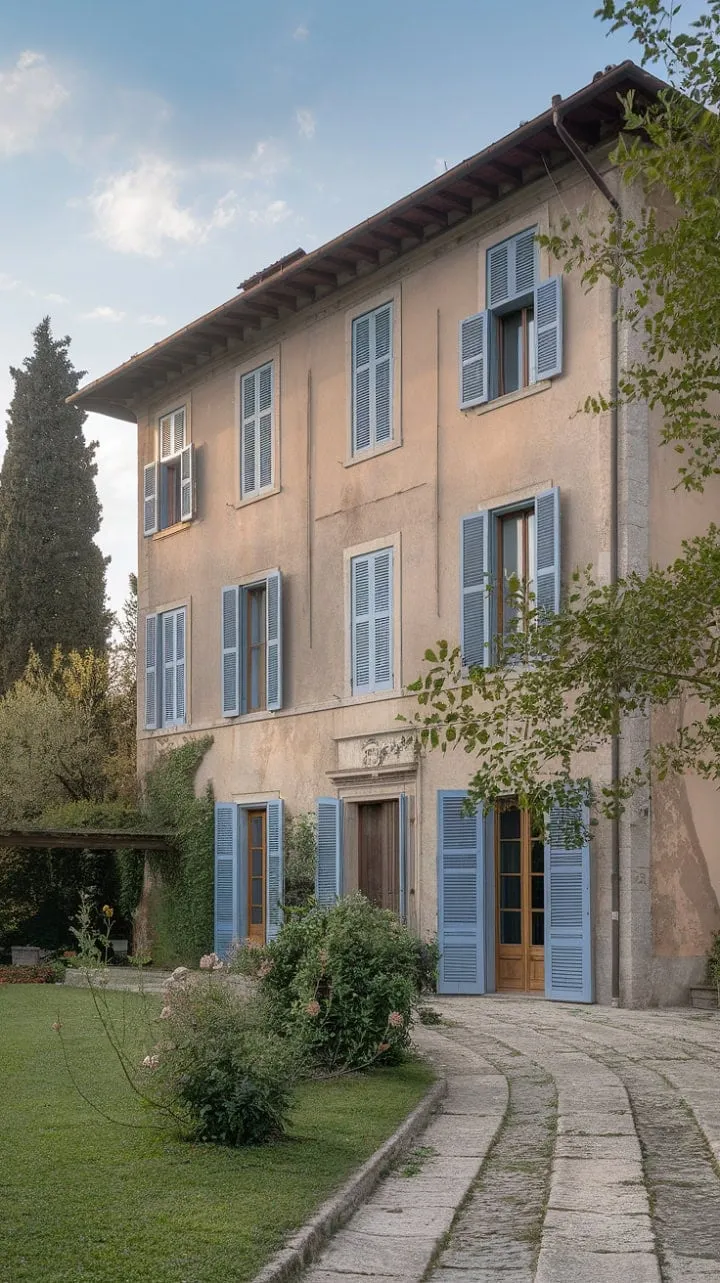 Bathed in sunlight, this serene Northern Italian villa with its terracotta roof and blue shutters invites leisurely strolls through lush gardens, embodying the essence of a perfect summer escape.