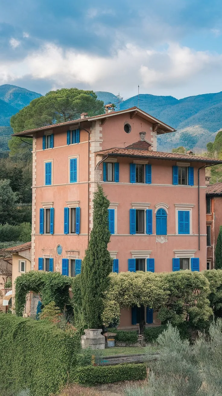 Northern Italy's picturesque villa, with its red roof, blue shutters, and arched windows, is nestled amongst vibrant greenery and majestic mountains.
