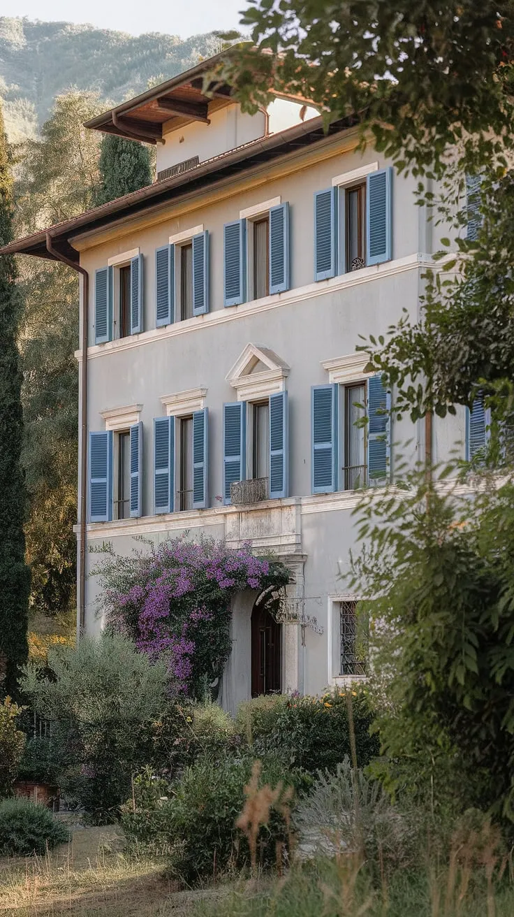 A sun-drenched Italian villa showcases charming blue shutters and lush greenery nestled in a mountainous landscape.