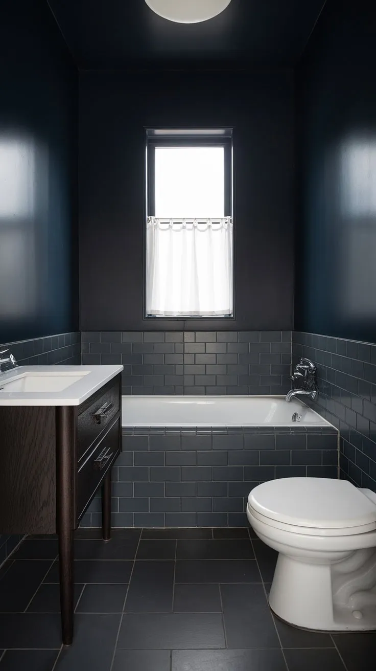 A sophisticated sanctuary. The deep navy walls and dark gray floor create a dramatic backdrop for the crisp white vanity and bathtub, a perfect blend of mid-century modern style and contemporary calm. A single window allows natural light to illuminate this small but perfectly formed space.