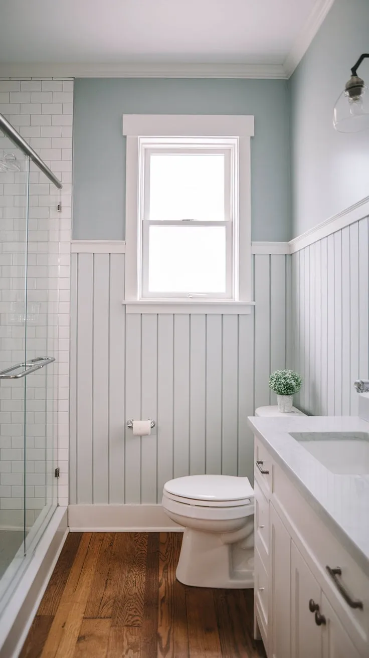 Clean lines and calming blues define this modern bathroom, where vertical shiplap wainscoting adds texture and visual interest, extending elegantly to the window for a seamless, sophisticated look. The light wood flooring complements the airy feel.